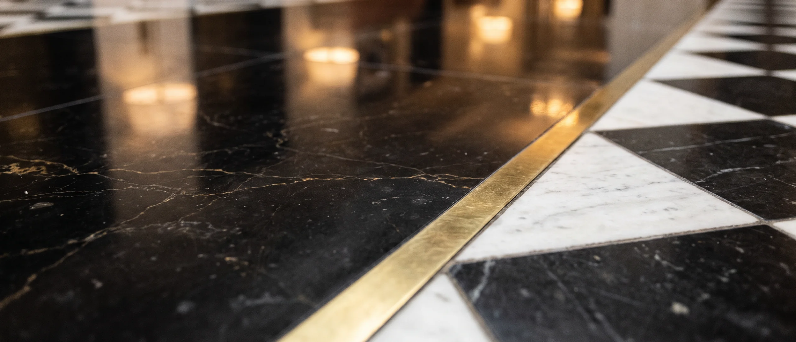 Polished black marble with a brass inlay strip meeting the edge of a black-and-white chequered marble floor, under soft reflected lamplight.